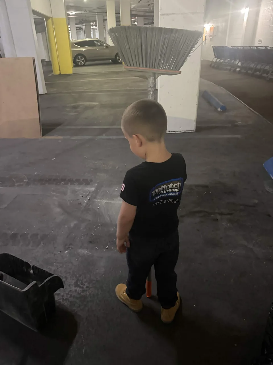 Jaxson, Justin and Katie's young son, in a Top Notch shirt holding a broom on a job site in a parking garage
