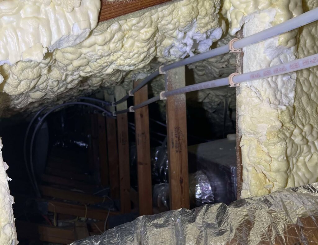 A view inside a dark attic showing thick, yellow spray foam insulation covering the rafters and ceiling deck. Several PEX water lines are routed horizontally and secured to wooden framing members, passing over a metallic flexible HVAC duct on the floor below.