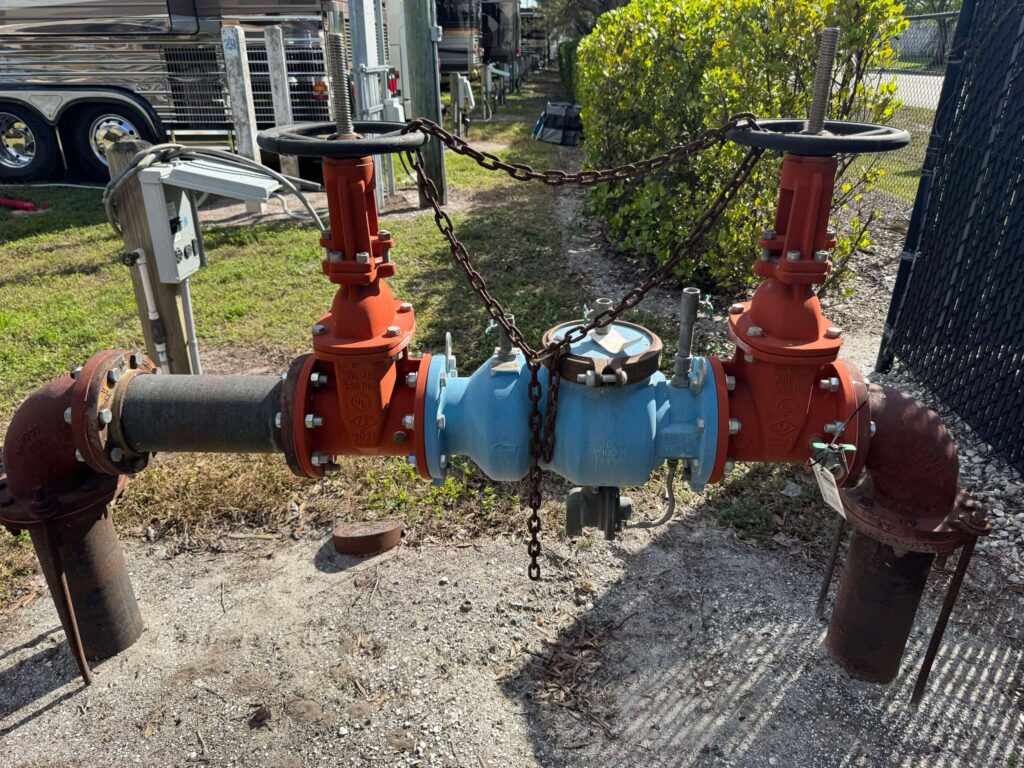 A large, outdoor water meter assembly consisting of a central blue meter and two large, vertical orange isolation valves on either side, all connected by horizontal pipes. A chain runs between the tops of the valves, securing the mechanism.