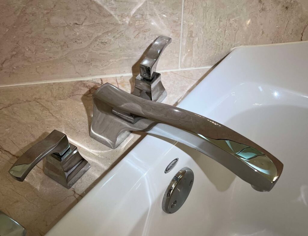 A close-up view of a modern, chrome, three-piece Roman tub faucet assembly mounted on the rim of a white bathtub. The spout has a long, slightly curved, flat design, and the two square-based handles have curved tops. The faucet is set against a tiled wall featuring pink and beige polished marble.