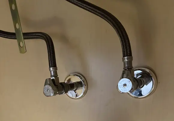 A close-up view of two modern, chrome-plated angle stop valves mounted on a beige wall under a sink. Each valve is connected to a faucet supply line covered in black braided stainless steel. A metal mounting bracket is visible in the upper left.