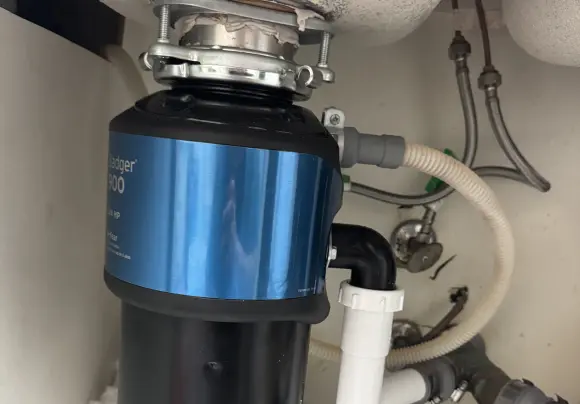 A close-up view underneath a kitchen sink showing a black and blue InSinkErator Badger 500 1/2 HP garbage disposal unit installed. The unit connects to the white drainpipe with a black elbow, and a flexible white hose, likely for a dishwasher drain, is connected to the side. Water supply lines are visible in the background.