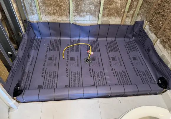 A bathroom area during construction shows a gray, rolled-out PVC shower pan liner installed over the floor base, ready for the mortar bed. A drain opening with a yellow wire is visible in the center, and the surrounding walls show exposed wooden studs and rough insulation.