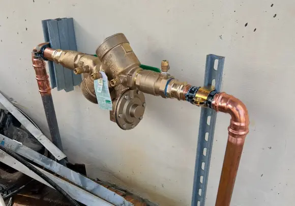 A close-up view underneath a sink showing a white drainpipe connected to a chrome-plated metal p-trap assembly. Heavy corrosion with light green and white residue is visible on the metal pipe, especially at the bend. Black putty or sealant is used around the joint where the metal pipe meets the white pipe, and a gray wire is draped across the pipes.
