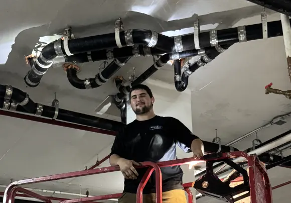 A smiling man in a dark work shirt, identified as a plumber by his uniform, is standing on a scissor lift platform looking up at a complex network of newly installed black drain or sewer pipes suspended from a concrete ceiling. The pipes are joined by couplings and held up by metal hangers.