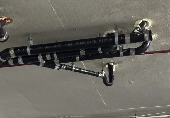 A view of plumbing exposed within a metal wall cavity showing a large black cast iron drain pipe on the left, connected to a horizontal copper drain line. White PEX pipes and copper water lines are visible to the right.