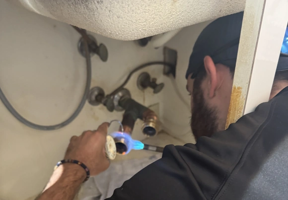 A close-up, over-the-shoulder view of a plumber working underneath a sink area. The plumber is wearing a black shirt and a baseball cap, holding a torch to heat a brass pipe fitting for soldering near the stub-out valves in the wall