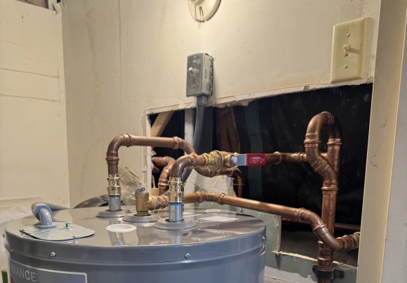 A close-up view of the top of a gray water heater tank with complex, newly installed copper piping connected to the inlet and outlet ports. A brass ball valve with a red handle is visible on the hot water line. The piping runs into an opening in the wall behind the heater, which shows damaged drywall and exposed wooden framing and insulation.