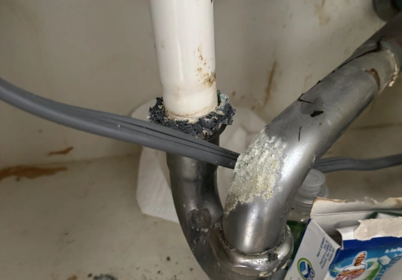 A close-up view underneath a sink showing a white drainpipe connected to a chrome-plated metal p-trap assembly. Heavy corrosion with light green and white residue is visible on the metal pipe, especially at the bend. Black putty or sealant is used around the joint where the metal pipe meets the white pipe, and a gray wire is draped across the pipes.