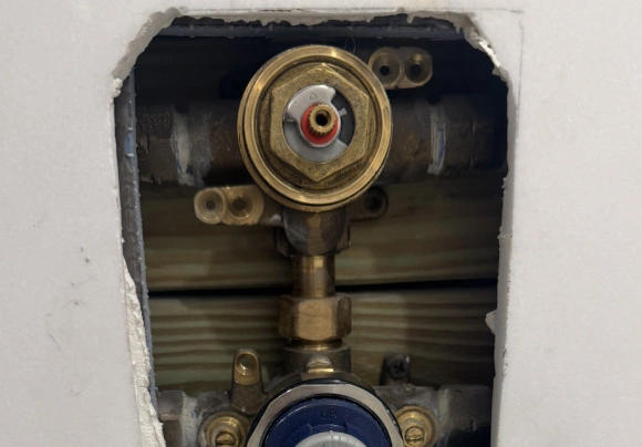 A close-up view of a partially installed brass shower or faucet valve cartridge mechanism exposed through a rectangular cut-out in drywall. The main valve body is centered, showing the brass retaining nut and a temperature limit stop or gear. Wooden wall framing is visible behind the piping.