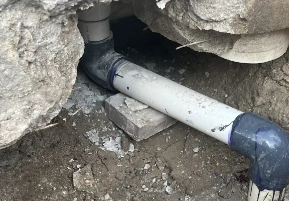 A close-up view of a newly installed section of white PVC drainpipe lying in a trench of dirt and small concrete fragments. The pipe is supported by a broken cinder block. It connects to a dark gray PVC elbow and fitting on either end, with visible purple primer/cement residue around the joints. The pipe runs through a gap in a broken concrete structure.