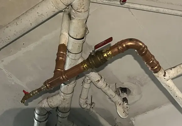 A dark attic space shows the roof deck and rafters covered in thick, heavily textured, yellowish-white spray foam insulation. Several white PEX water lines are secured horizontally to exposed vertical wooden framing members. A large, silver, flexible HVAC duct is partially visible on the floor below.